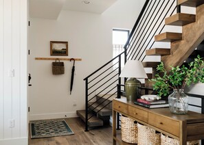 A staircase with wooden steps and black railings, a wooden console table with a lamp, a vase with plants, and a woven basket on the floor. - Luxury 5BR with hot tub, heated game garage, mini golf, outdoor projector (Denver)