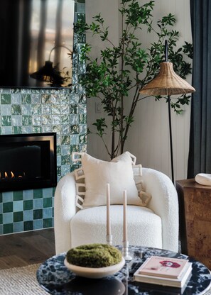 A cozy living room with a fireplace, a white armchair, a floor lamp, and a small table with a book and candles. - Luxury 5BR with hot tub, heated game garage, mini golf, outdoor projector (Denver)