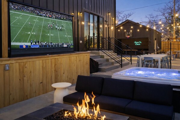 An outdoor seating area with a fire feature, a large screen displaying a football game, and a building with a neon sign. - Luxury 5BR with hot tub, heated game garage, mini golf, outdoor projector (Denver)