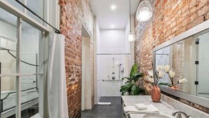 Un baño moderno con pared de ladrillo, un espejo grande, ducha y lavamanos con un florero.