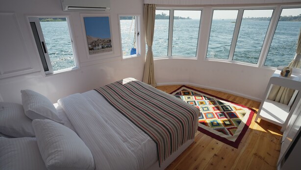 Een slaapkamer met een bed, een raam met uitzicht op zee, een klein raam met een afbeelding en een kleurrijk vloerkleed.