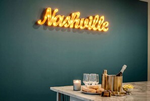 Eine Neonreklame mit dem Schriftzug 'Nashville' an einer türkisblauen Wand, eine Marmor-Bar mit Gläsern, einer Kerze und Champagnerflaschen.