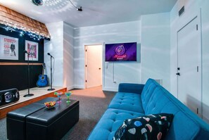 Ein modernes Wohnzimmer mit einem blauen Sofa, einem schwarzen Hocker, einer Gitarre und einem Neon-Schild, das eine Karaoke-Veranstaltung anzeigt.