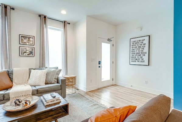 A modern living room with a sofa, a coffee table with books and a tray, a door, and a wall with a framed quote. - Summer rooftop decks, karaoke room, wet bar near Broadway and Gulch (Nashville)