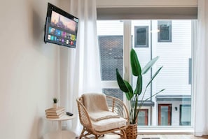 A living room with a mounted TV, a wicker chair, a small table with books and a plant, and a view of a building through large windows. - Summer rooftop decks, karaoke room, wet bar near Broadway and Gulch (Nashville)