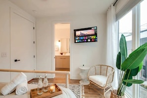 A modern hotel room with a wooden floor, a white bed with a white pillow, a wicker chair, a large green plant, a small round table with a candle, and a TV mounted on the wall displaying a cityscape. - Summer rooftop decks, karaoke room, wet bar near Broadway and Gulch (Nashville)