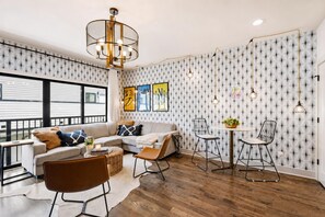Un salon moderne avec un canapé d’angle, une table à manger avec des chaises et une grande fenêtre équipée de stores.