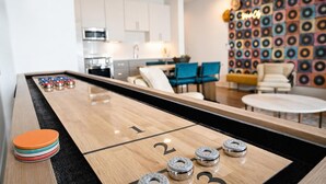 Ett shuffleboard-bord med numrerade markeringar och puckar, en hög med färgglada puckar och ett modernt vardagsrum med en vägg med mönster och möbler.