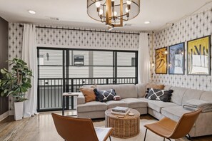 A modern living room with a sectional sofa, a round wicker coffee table, a potted plant, and a large window with a view of a building. - Fall getaway near Belmont & Hillsboro Village, Nashville, sleeps 12 (Nashville)