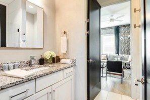 A modern bathroom with a large mirror, white cabinets, and a granite countertop. There is a towel rack with a white towel and a vase with flowers. The bathroom leads to a living area with a black chair and a sofa. - Summer rooftop deck, ping pong lounge, skyline views near Broadway (Nashville)
