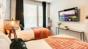 Habitación de hotel con dos camas, un televisor en la pared mostrando un paisaje urbano, una mesita con un jarrón y una ventana con persianas.