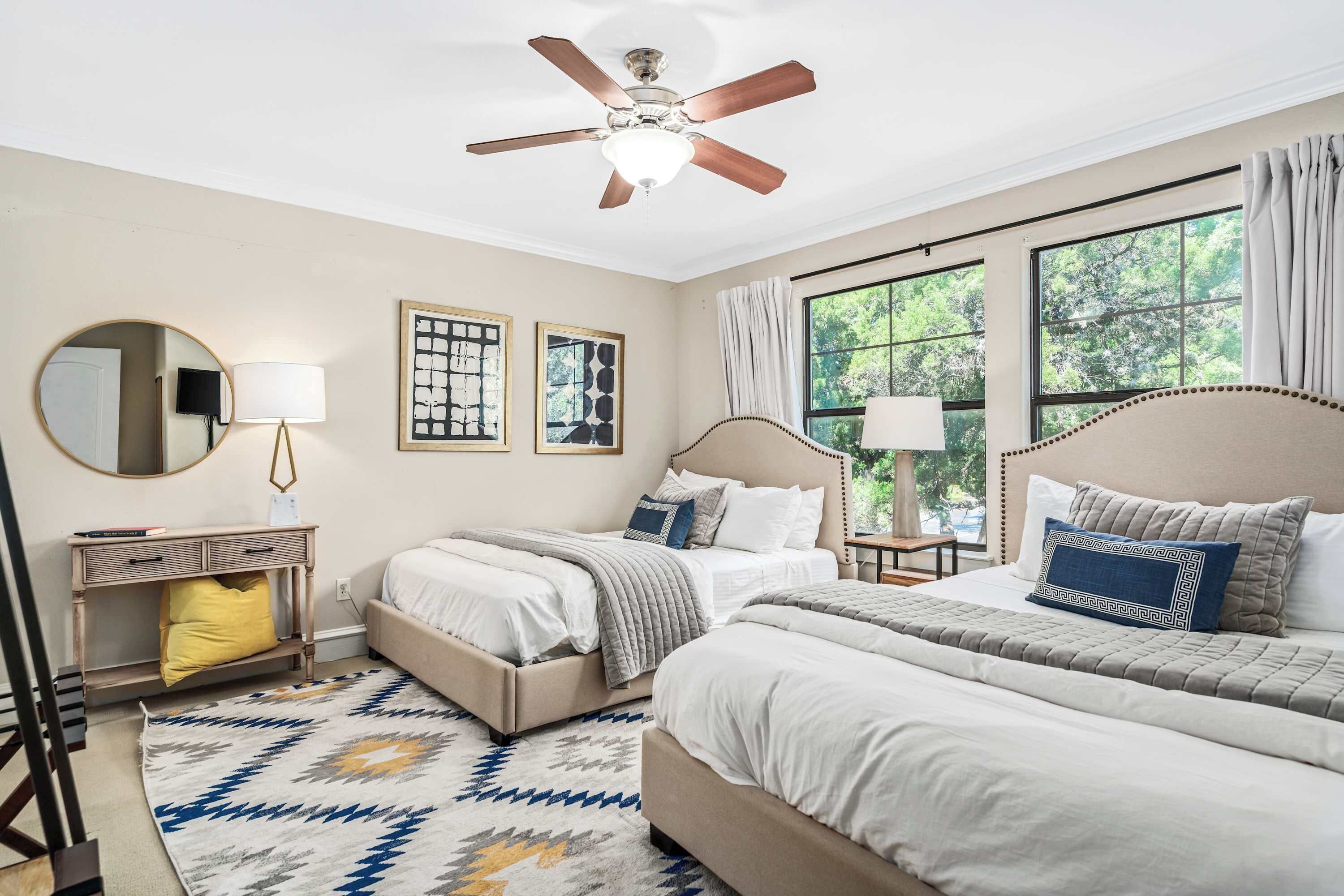 A bedroom with two beds, a ceiling fan, a large window with curtains, a bedside table with a lamp, and a rug.
