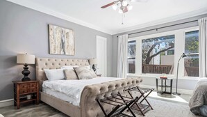 A bedroom with a tufted headboard bed, a nightstand with drawers, a floor lamp, and a large window with a view of trees. - ATX Oasis / Pool / Firepit / Billiards (Austin)