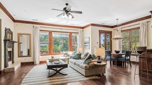 A spacious living room with hardwood floors, a fireplace, a ceiling fan, and large windows offering a view of the outdoors. - ATX Oasis / Pool / Firepit / Billiards (Austin)
