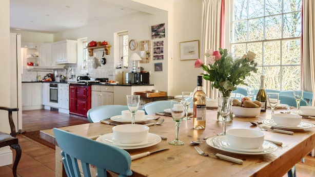 Un coin repas avec une table en bois mise pour quatre personnes, des assiettes blanches, des verres et des couverts. Un vase rempli de fleurs est posé sur la table.