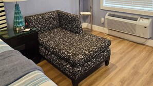 Een slaapkamer met een gestreept sofa, een nachtkastje met een lamp, een staande lamp, een raam met zonwering en een airconditioner.