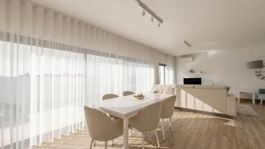 A modern dining area with a white table and chairs, a kitchen in the background, and large windows with sheer curtains.