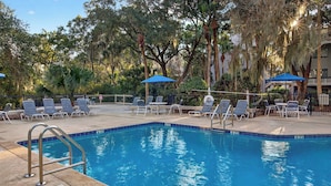 A swimming pool with lounge chairs and umbrellas, surrounded by trees and a building. - Treetops 4904 by Avantstay Hilton Head Condo + Balcony + Shared Pool + Tennis Court + Near Beach and Dining (Hilton Head Island)