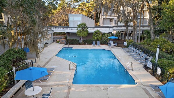 A rectangular pool with a white deck, surrounded by lounge chairs and umbrellas, with a bridge and trees in the background. - Treetops 4904 by Avantstay Hilton Head Condo + Balcony + Shared Pool + Tennis Court + Near Beach and Dining (Hilton Head Island)