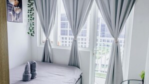 Een slaapkamer met een bed, grijze gordijnen, een raam met uitzicht op gebouwen en een kleine tafel met een plant.