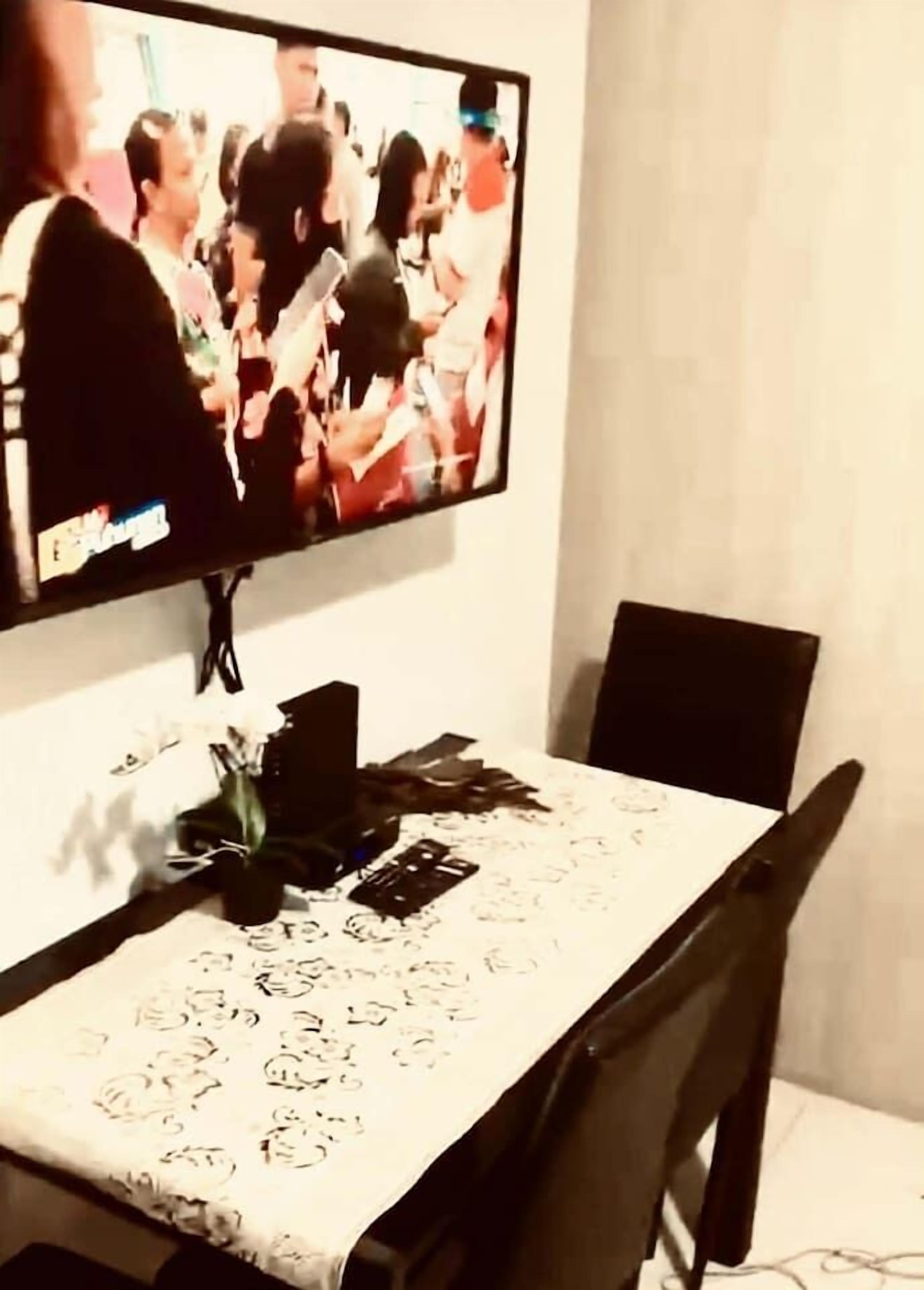Un comedor con televisión instalada en la pared mostrando un programa, una mesa con mantel floral y un jarrón con flores.