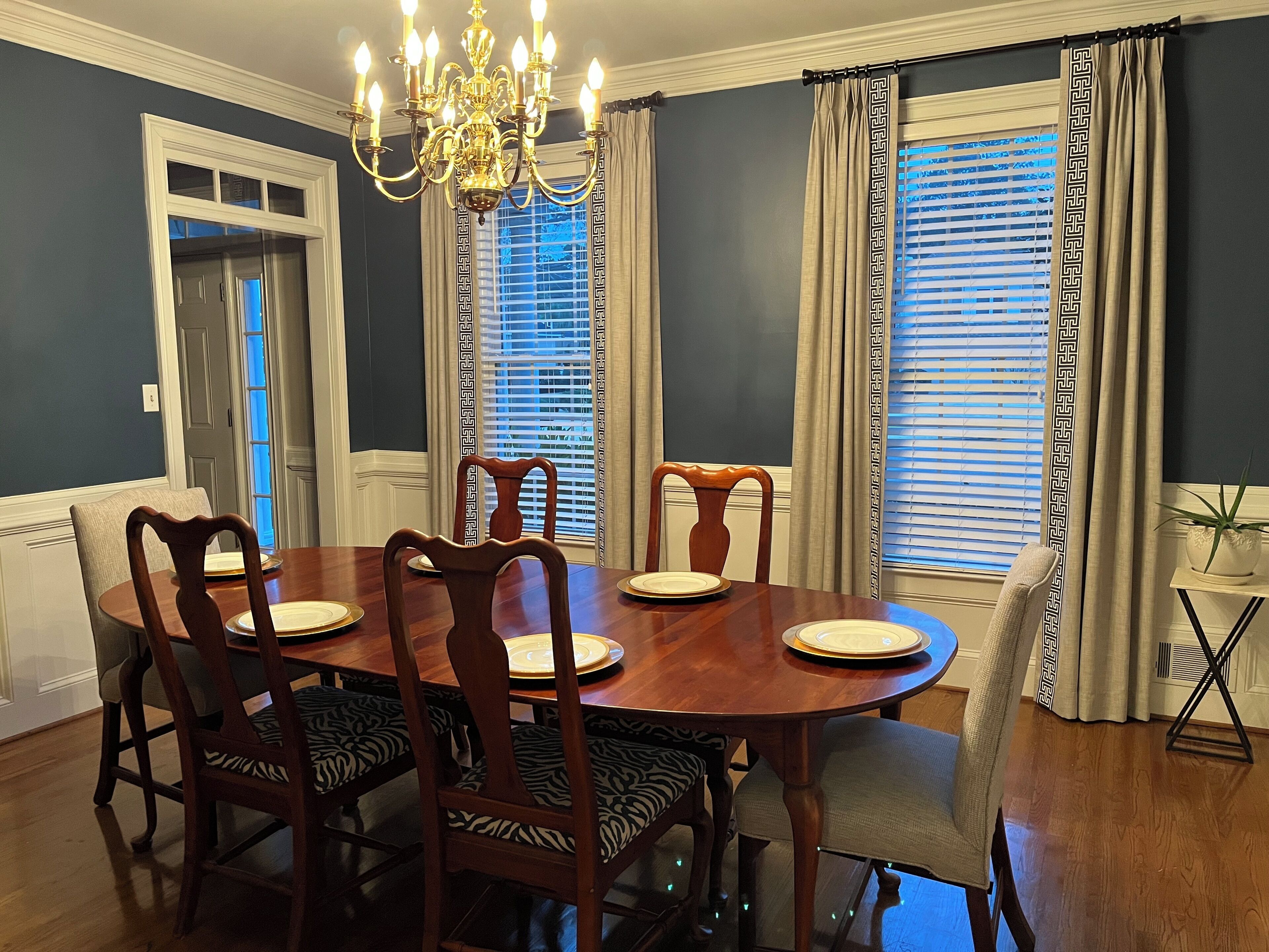 A dining room with a round table, six chairs, a chandelier, and windows with patterned curtains.