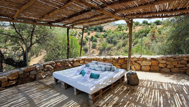 A bed with towels and a glass of wine on a wooden platform, surrounded by stone walls and a thatched roof, overlooking a hilly landscape. - Refugio La Sima El Gastor by Ruralidays (Cádiz)