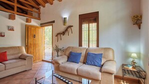A living room with two beige sofas, one with a red pillow, and a small table with a lamp and a book. There is a wooden door, a window with a view, and a wall-mounted decoration. - Refugio La Sima El Gastor by Ruralidays (Cádiz)