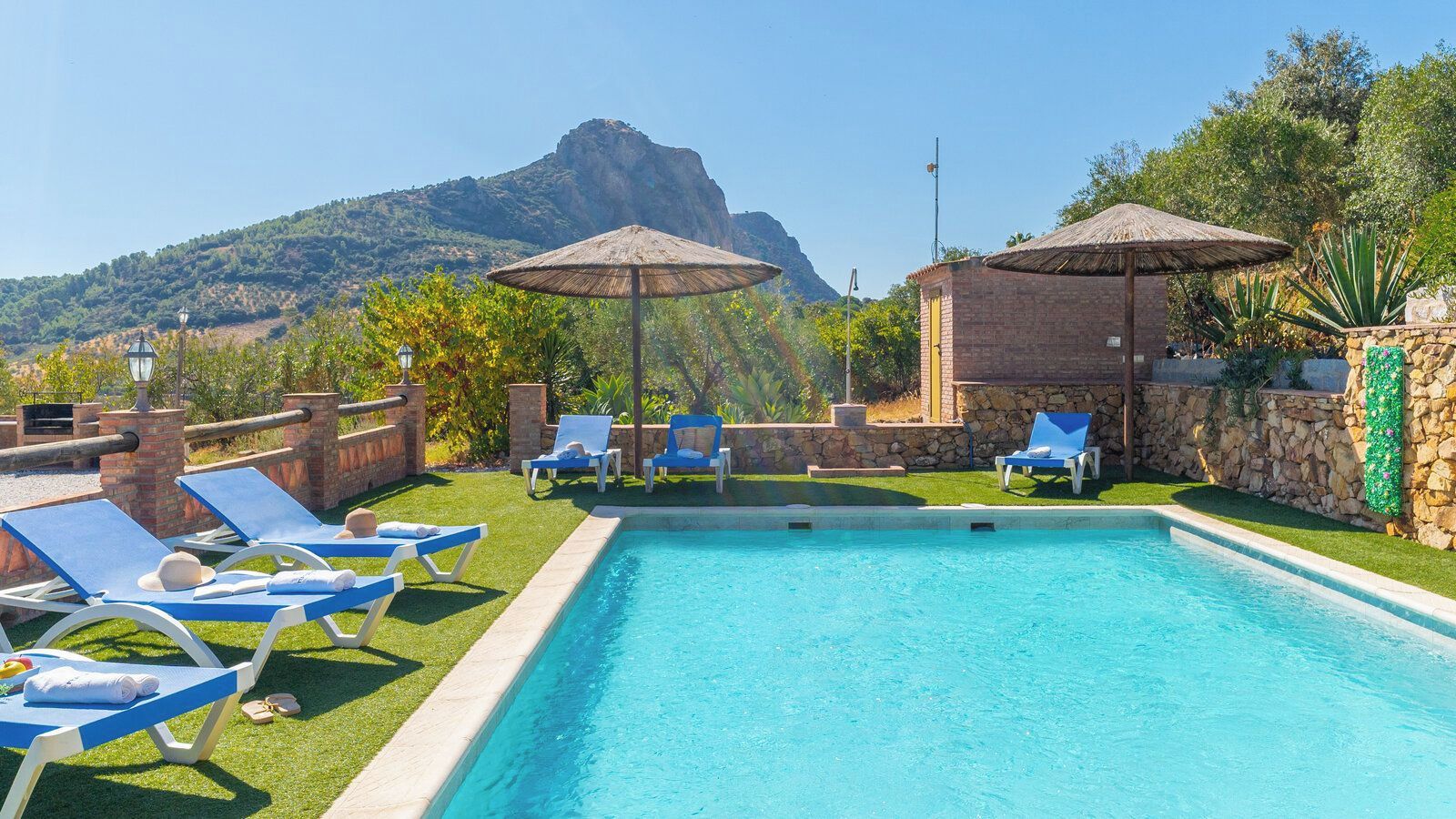A swimming pool with lounge chairs and umbrellas, surrounded by a stone wall and greenery, with a mountain in the background.