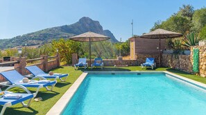 A swimming pool with lounge chairs and umbrellas, surrounded by a stone wall and greenery, with a mountain in the background.