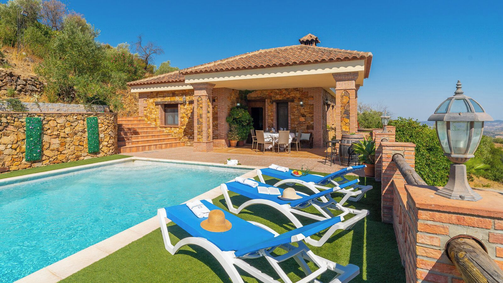 Un área junto a la piscina con sillones reclinables, un edificio de piedra con techo de tejas y un cielo azul despejado.