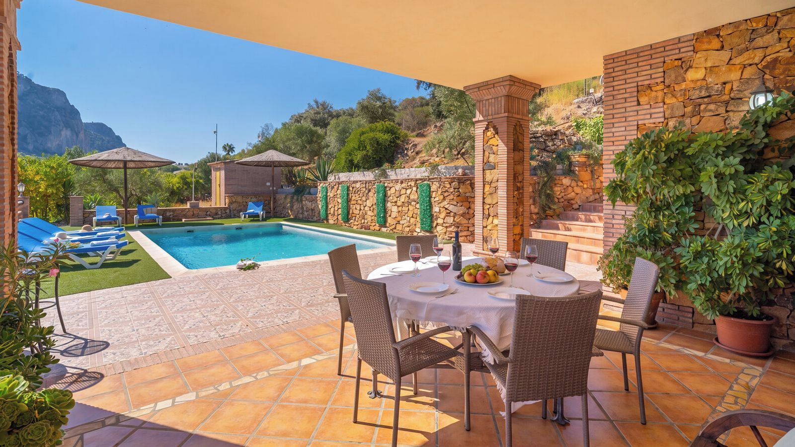 A poolside area with a table set for dining, lounge chairs, and a stone wall.