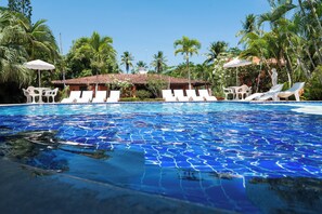 Ein Pool mit klarem, blauem Wasser, umgeben von weißen Liegestühlen und Sonnenschirmen, mit Palmen und einem Gebäude im Hintergrund.