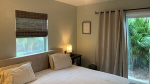 Ein Schlafzimmer mit Bett, Deckenventilator, einem Fenster mit Jalousien, einem Nachttisch mit Lampe und Blick ins Grüne.