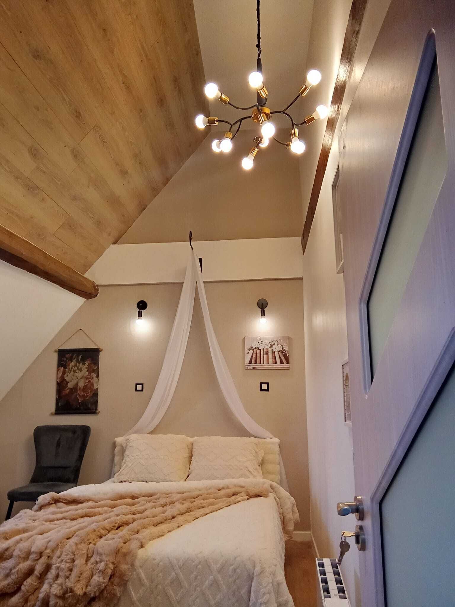 Ett sovrum med ett träpanelat tak, en säng med vita sängkläder, en sänghimmel, en stol och en väggmonterad lampa.