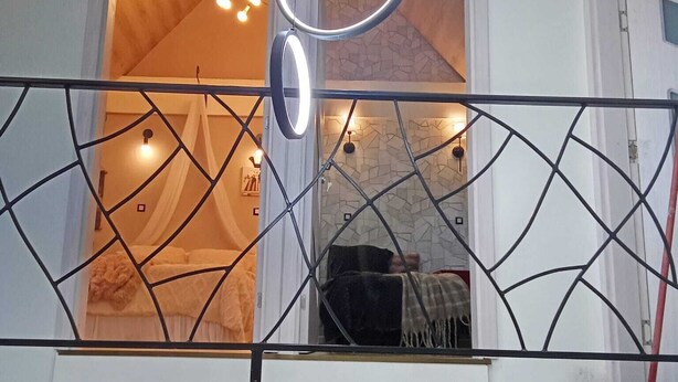 A balcony with a geometric metal railing, a hanging light fixture, and a view into a room with a bed and a fireplace. - Maison moderne à 20 minutes de Strasbourg (Kurtzenhouse)