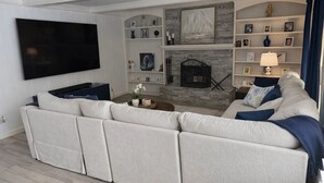 A living room with a large sectional sofa, a flat-screen TV, a fireplace, and shelves with framed pictures and decorative items. - Marvelous 4-bedroom house with Pool in lovely Lenexa World Cup Ready (Lenexa)