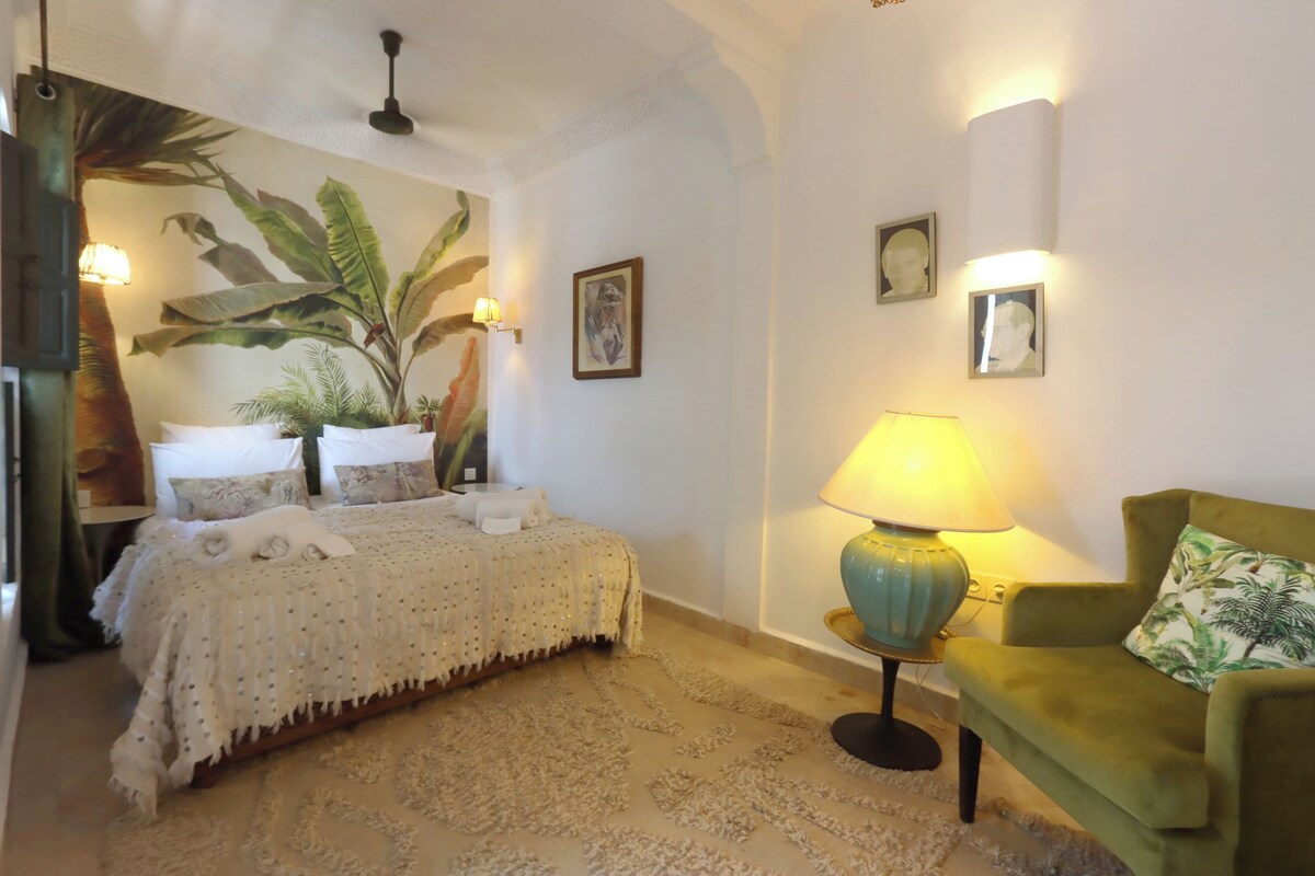 A room with a bed, a green armchair, a lamp, and a large plant.