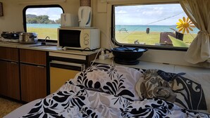 Eine kompakte Küchenzeile mit Mikrowelle, Herd und Spüle. Im Vordergrund steht ein Bett mit gemusterten Betttextilien, und ein Fenster bietet Blick auf Strand und Meer.