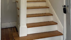 Una escalera con peldaños pintados de blanco y barandillas de madera, que lleva a una puerta con pomo negro.