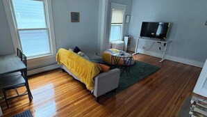 Un espacio habitable compacto con una cama, una mesita, un televisor y una ventana.