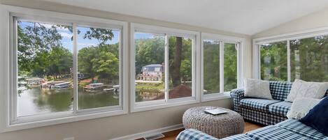 Ein Zimmer mit großen Fenstern mit Blick auf einen See und ein Haus. Es gibt ein kariertes Sofa und einen kleinen Hocker.