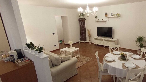 Ein Wohnzimmer mit Fernseher, Sofa, Esstisch und Stühlen. Es gibt einen Kamin, eine Pendelleuchte und Zimmerpflanzen.