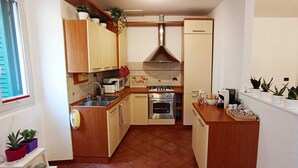 Eine kleine Küche mit Holzschränken, einem Herd, einer Mikrowelle und einem Spülbecken. Auf den Arbeitsflächen stehen Blumentöpfe und es gibt ein Fenster mit Jalousien.