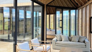 A modern interior with a high ceiling, wooden beams, and large glass windows offering views of the outdoors.