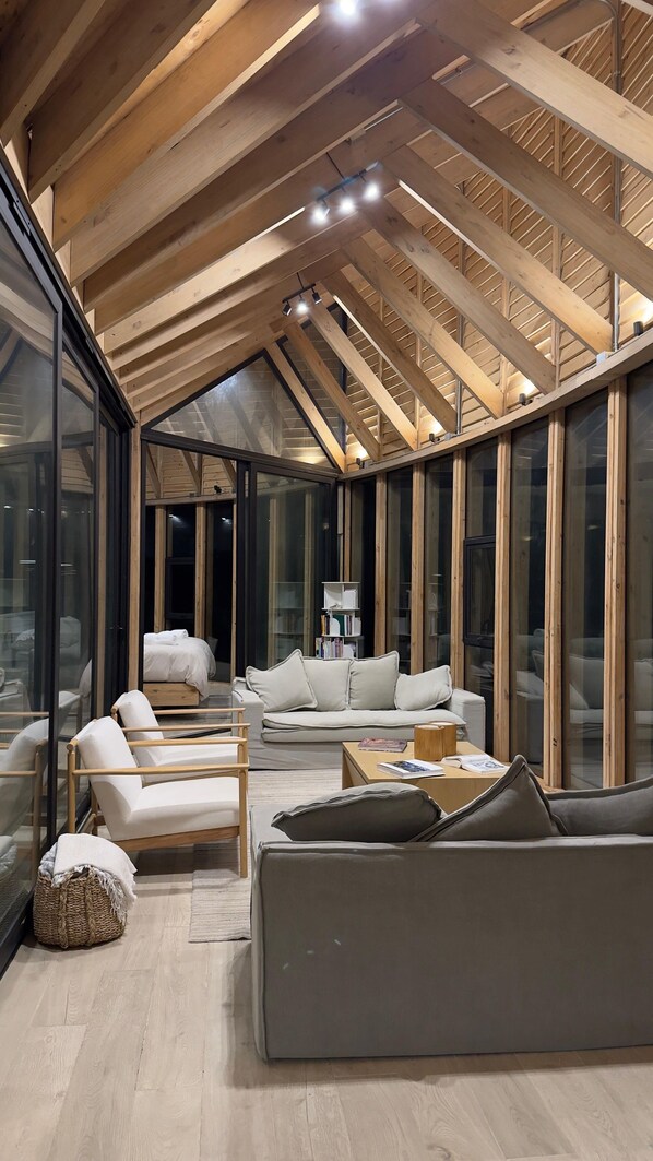A modern interior with a high ceiling, wooden beams, and large glass windows. There is a sofa, a chair, and a small table with books.