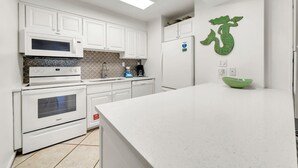 A modern kitchen with white cabinetry, a microwave, an oven, a refrigerator, and a green decorative mermaid wall art. - Jetty East 106b (Destin)