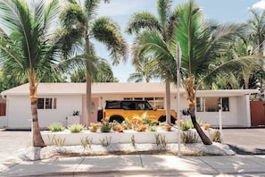 Una casa blanca con un SUV amarillo estacionado en la entrada, rodeada de palmeras y un jardín bien cuidado.