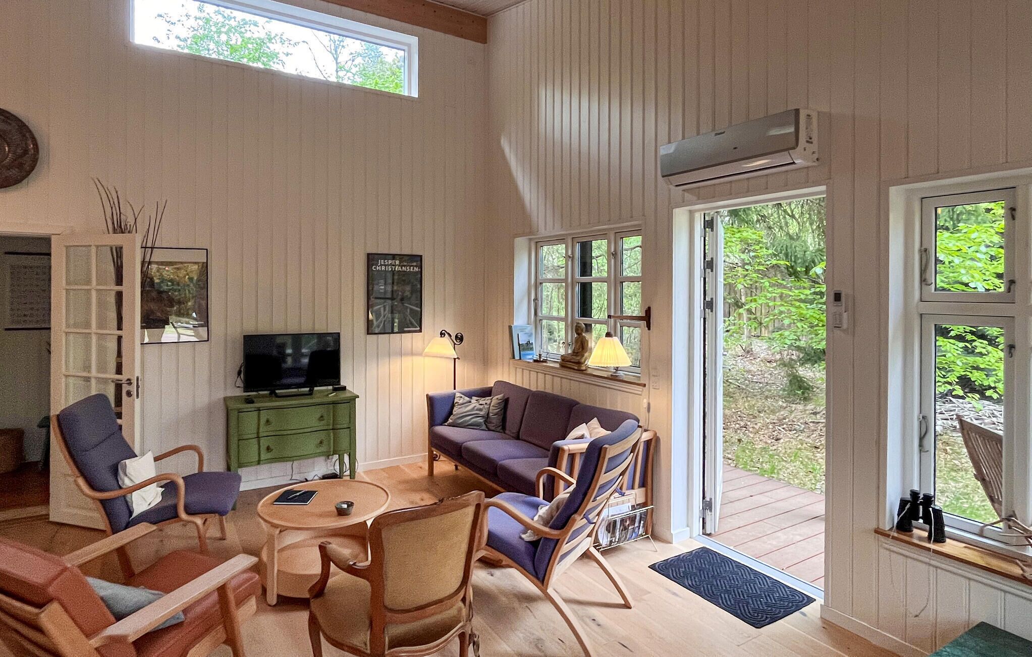En hyggelig stue med fjernsyn, et grønt skab, en lilla sofa, træstole og store vinduer.