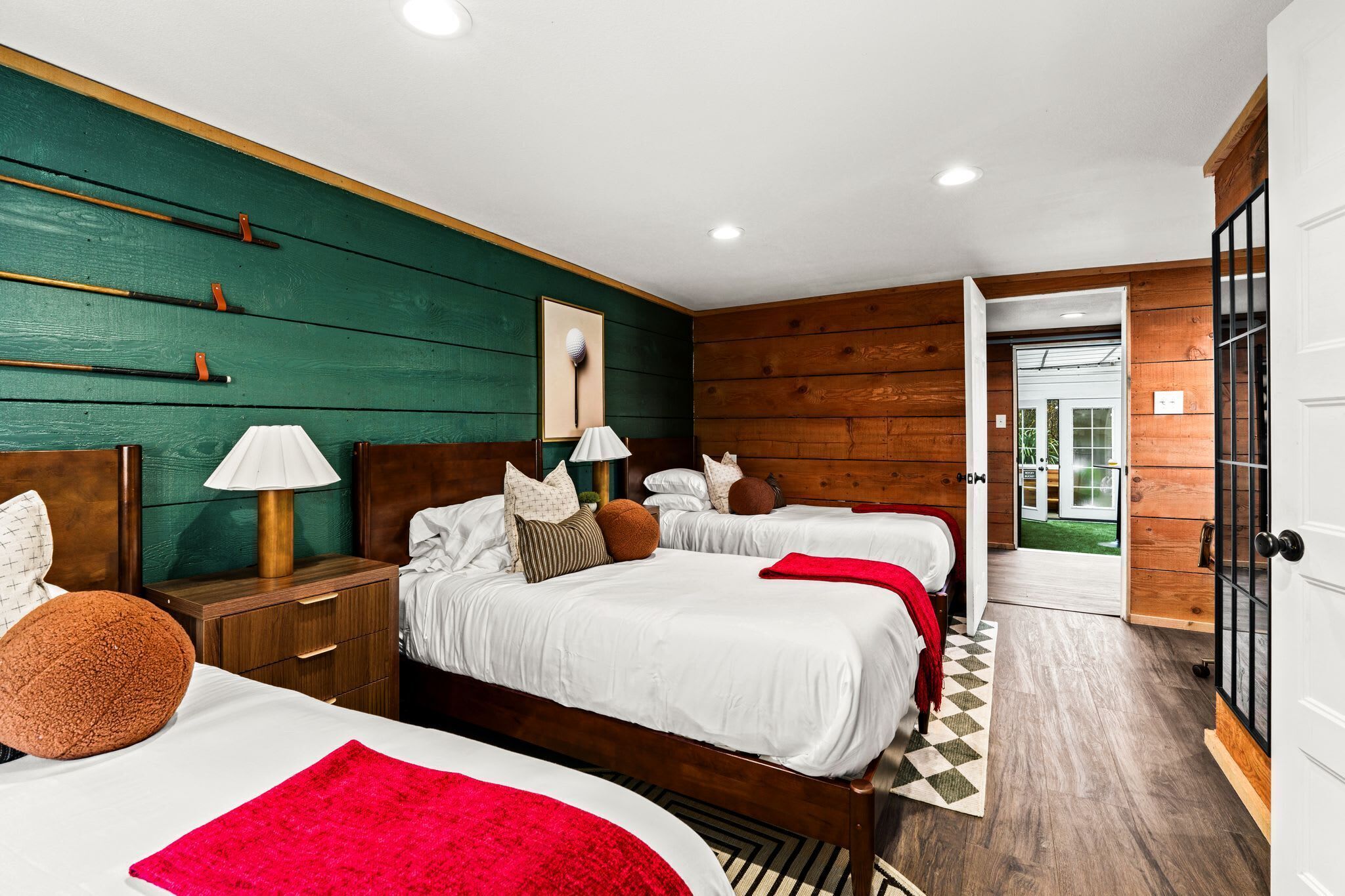 A hotel room with two beds, a wooden headboard, a nightstand with a lamp, and a view of the outdoors through an open door.